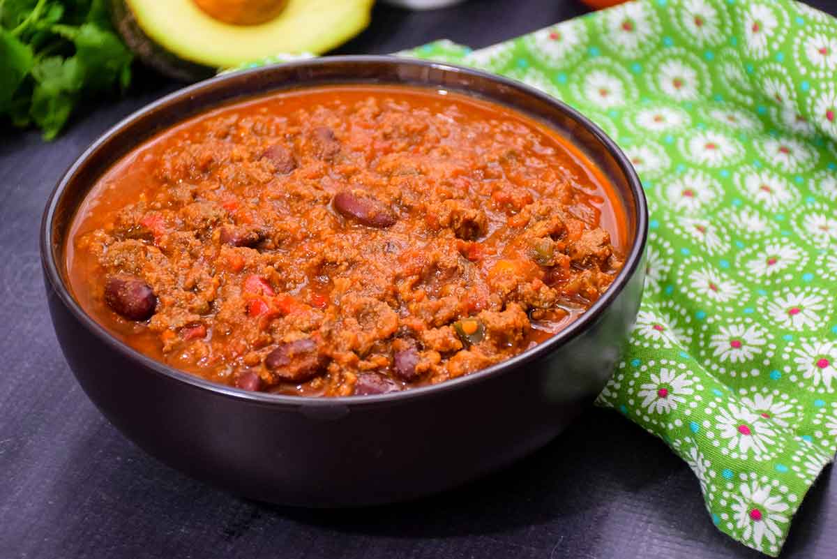 A close up of a bowl filled with chili made with ground venison and beans, with an chopped avocado in a white bowl in the back ground and an avocado half next to it.