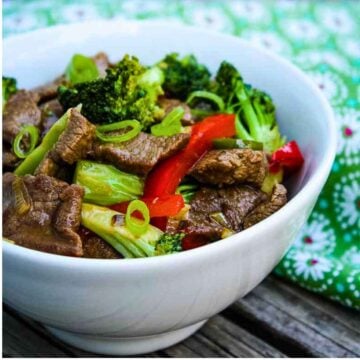 Bowl of venison stir fry with broccoli, red bell pepper, and scallions, served on a rustic wooden surface with a green floral napkin.