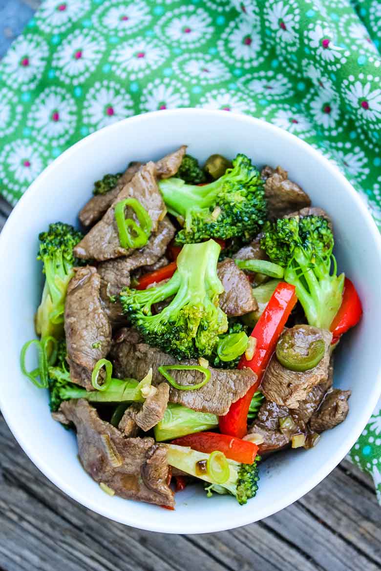 Overhead view of a bowl of venison stir fry with bright green broccoli, red bell peppers, and scallions on a green floral cloth.