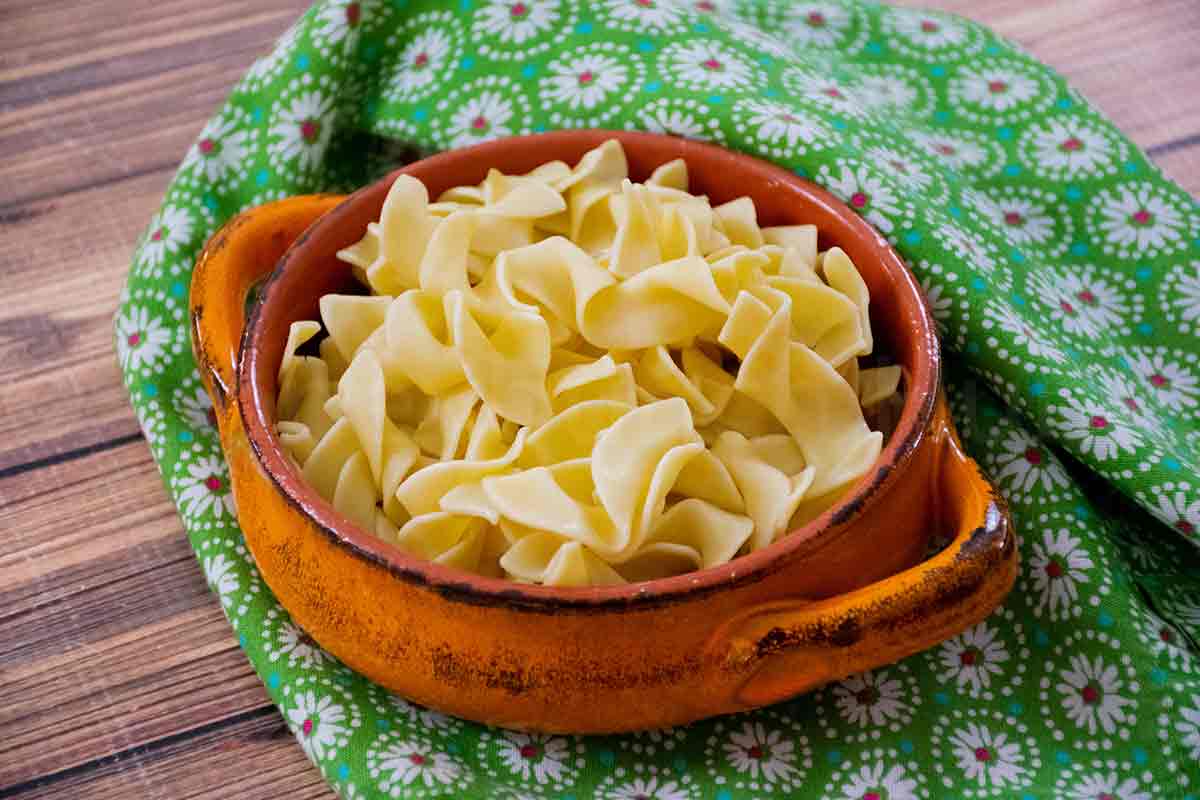 Bright orange bowl filled with cooked egg noodles placed on a rustic wooden surface with a green floral cloth in the background.