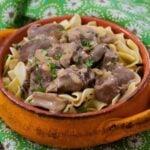 Close up of venison stroganoff served over egg noodles in a rustic orange bowl with green parsley garnish.
