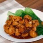 A white plate with Vietnamese caramel chicken, featuring glazed bite sized chicken pieces in a rich brown sauce, served alongside steamed baby bok choy on a checkered cloth napkin background
