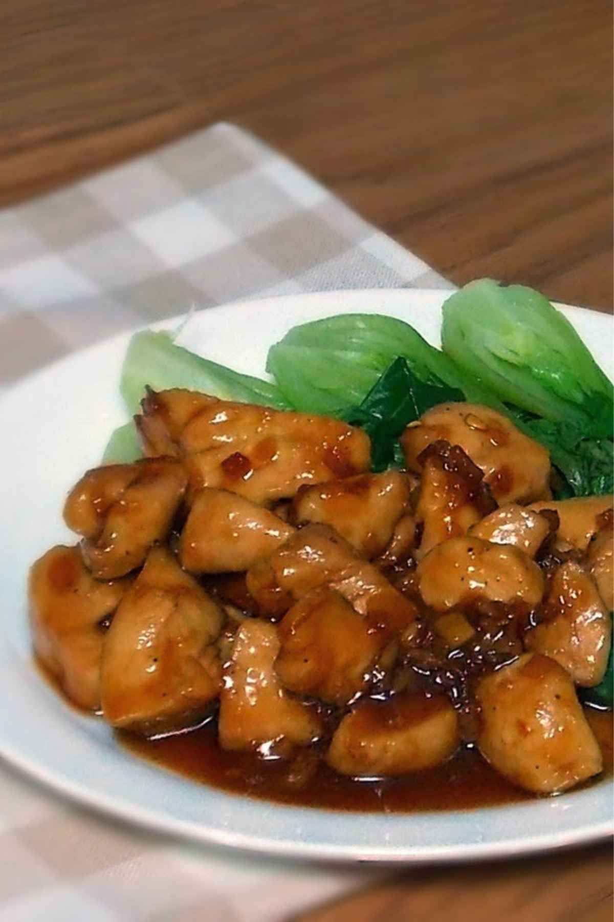 A white plate with Vietnamese caramel chicken, featuring glazed bite sized chicken pieces in a rich brown sauce, served alongside steamed baby bok choy on a checkered cloth napkin background