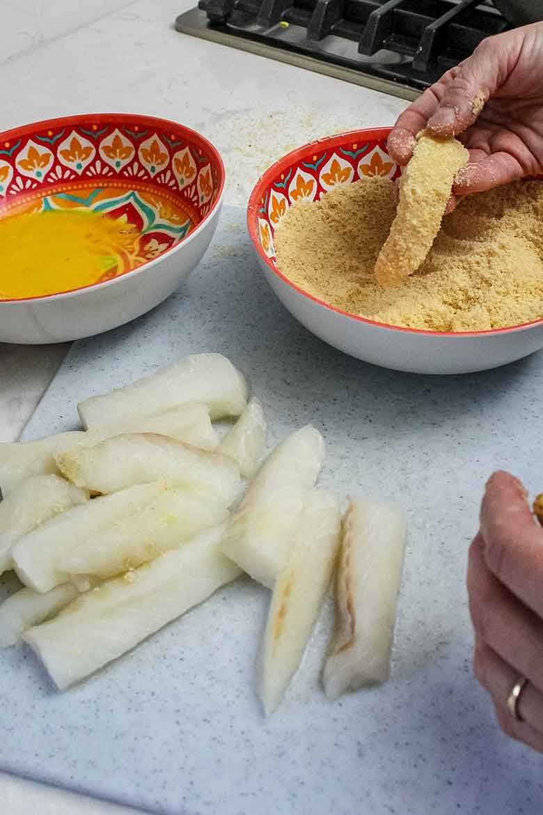 A hand dips a piece of cod into a bowl of breading mixture next to a bowl of beaten egg, with raw fish strips on a cutting board.