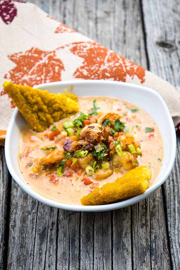 A bowl of creamy Cajun shrimp topped with chopped green onions and herbs garnished with crispy polenta triangles, photographed at an angle on a rustic wooden surface.