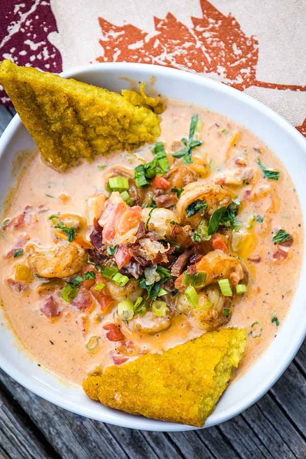 A close up top down view of a bowl of Cajun Shrimp in a creamy tomato-based sauce, garnished with scallions, parsley, and bacon bits, served with golden fried polenta wedges.