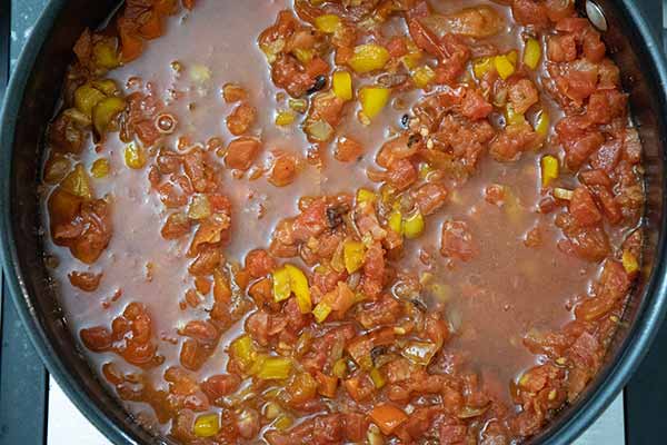 A close up of a skillet filled with diced tomatoes and yellow bell peppers simmering in a thin, brothy sauce, forming the tomato base for a spicy shrimp recipe.
