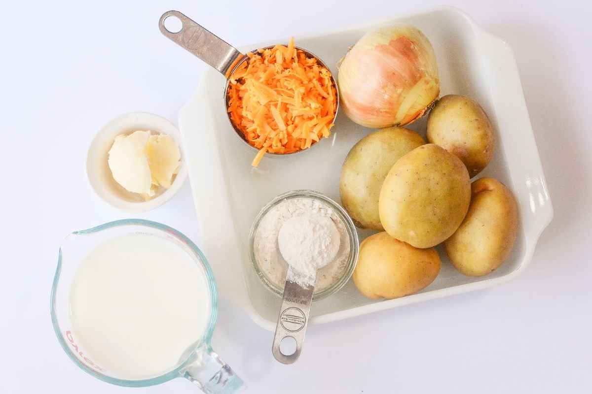A white dish with whole potatoes, onion, shredded cheddar cheese, flour, butter, and a measuring cup of milk on a white background.