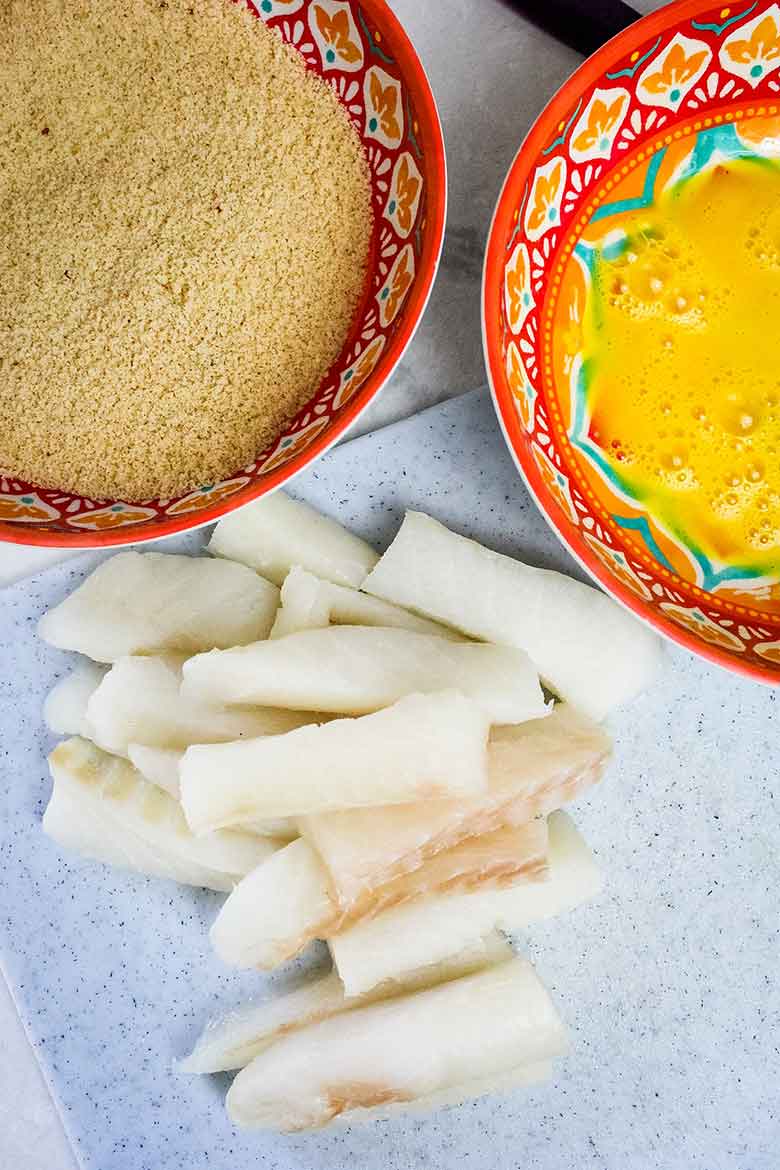 Overhead view of raw cod fish strips on a white cutting board beside bowls of breading and beaten egg mixture, ready for coating.