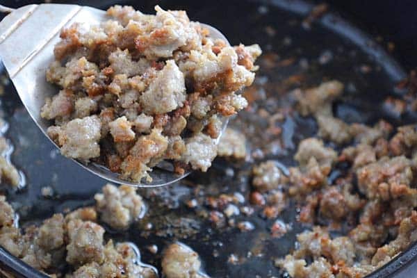 A metal spatula lifts browned, crumbled sausage from a skillet with visible oil.