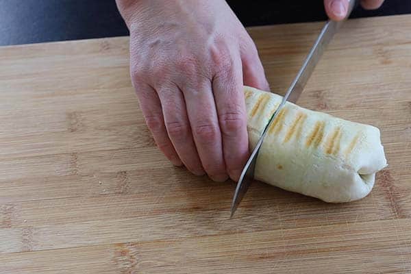 A hand cuts a grilled burrito in half on a wooden cutting board using a large knife.