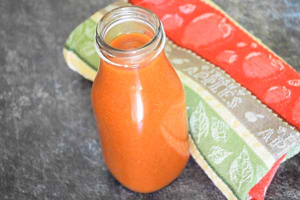 A glass bottle filled vibrant homemade taco sauce, set on a dark countertop with a colorful striped towel in the background.