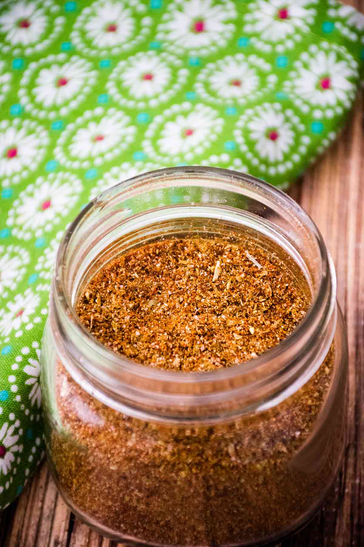 A glass jar filled with finely blended homemade taco seasoning, sitting on a wooden surface next to a bright green cloth decorated with white and red floral patterns.