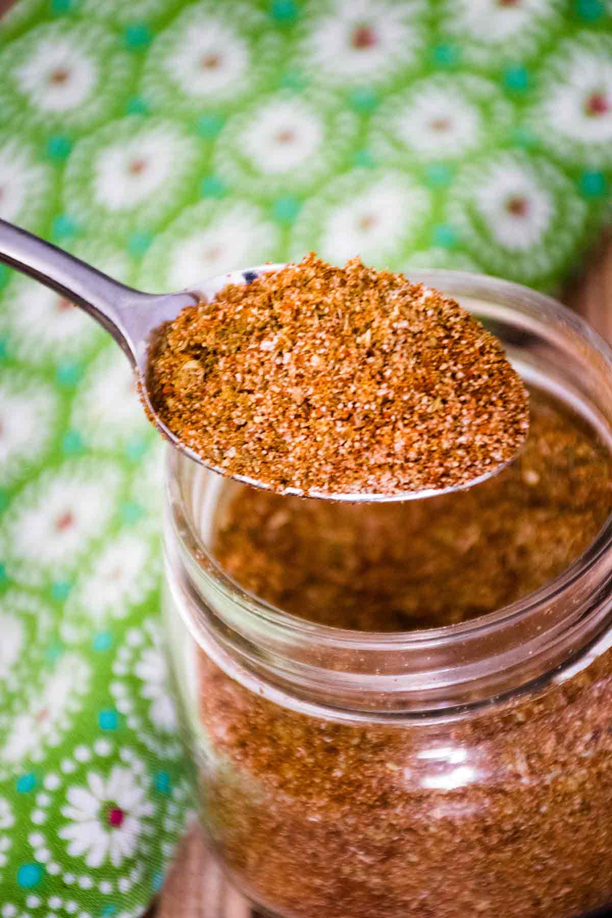 A spoonful of finely blended homemade taco seasoning held above a jar with a background of a green cloth patterend with white daisies.