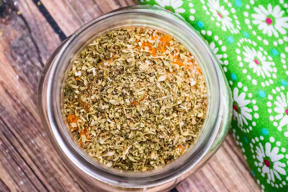 Top down view of a glass jar filled with a blend of dried herbs and spices on a wooden surface next to a green floral patterned cloth.