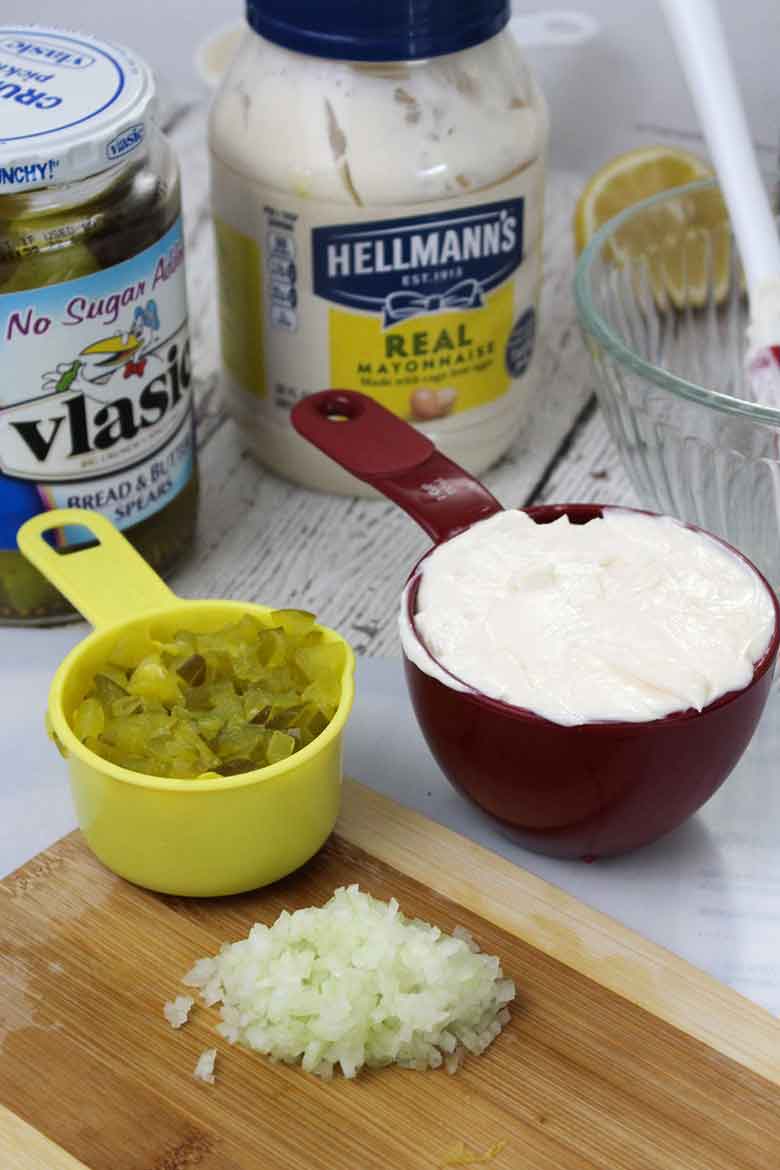 Ingredients for homemade tartar sauce on a cutting board, including chopped pickles in a yellow cup, mayonnaise in a red cup and finely diced onion, with jars of mayo and pickles in the background.