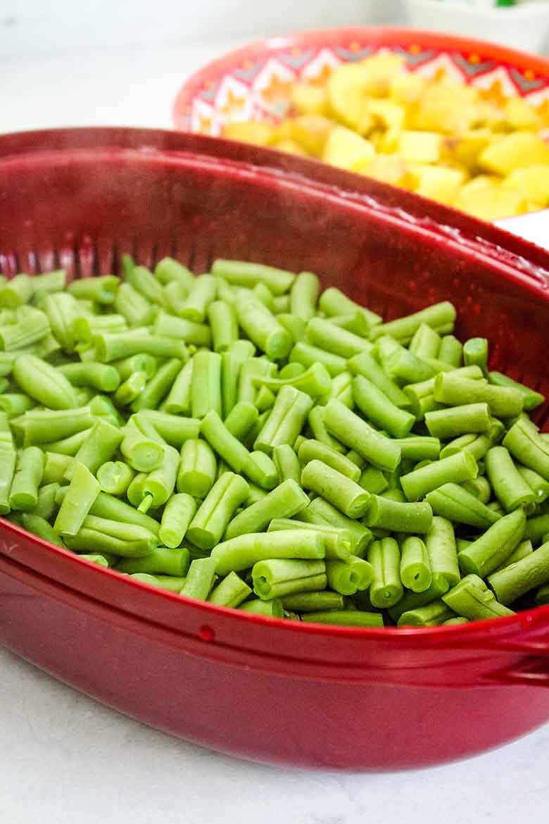 A red bowl filled with freshly steamed and chopped green beans, with a second bowl of cooked potatoes in the background.