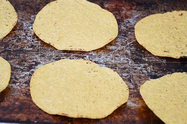 Several plain tostada shells arranged in rosw on a well used metal baking sheet.