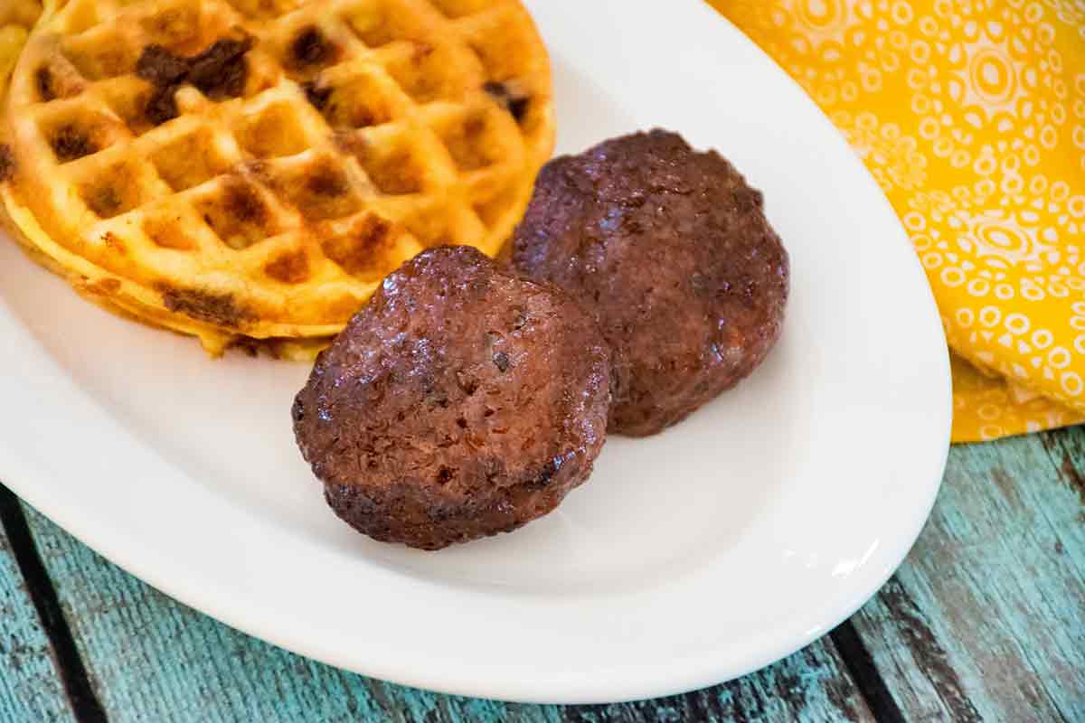Two cooked breakfast sausage patties on a white plate with a chocolate chip waffle next to it.