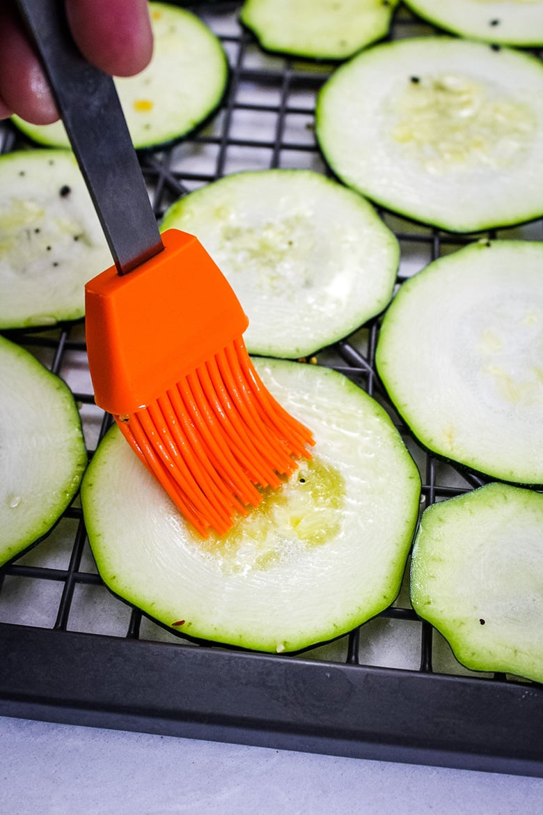 A silicone brush spreading olive oil on raw zucchini slices before seasoning and dehydrating.