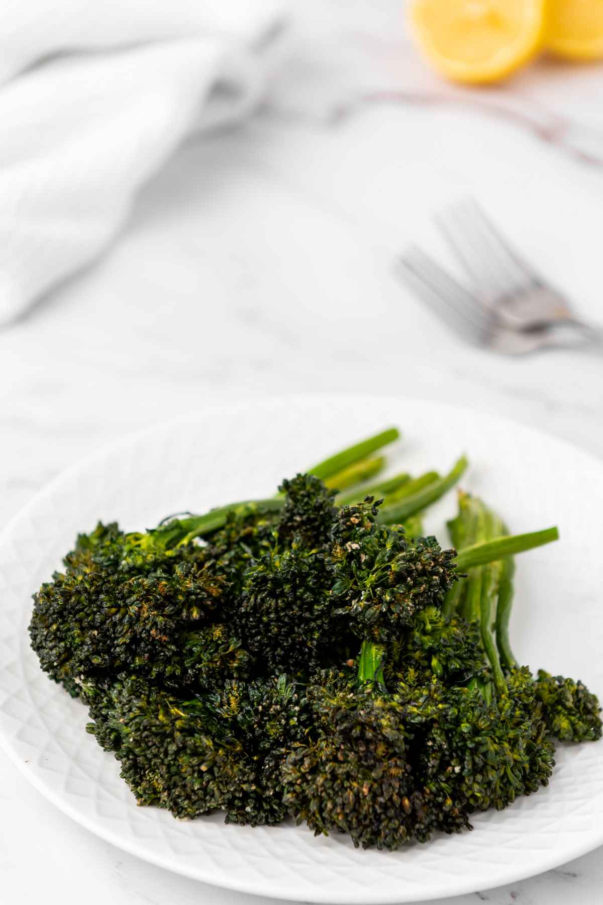 Plated air fryer broccolini on a white marble surface with lemon wedges and forks in the background