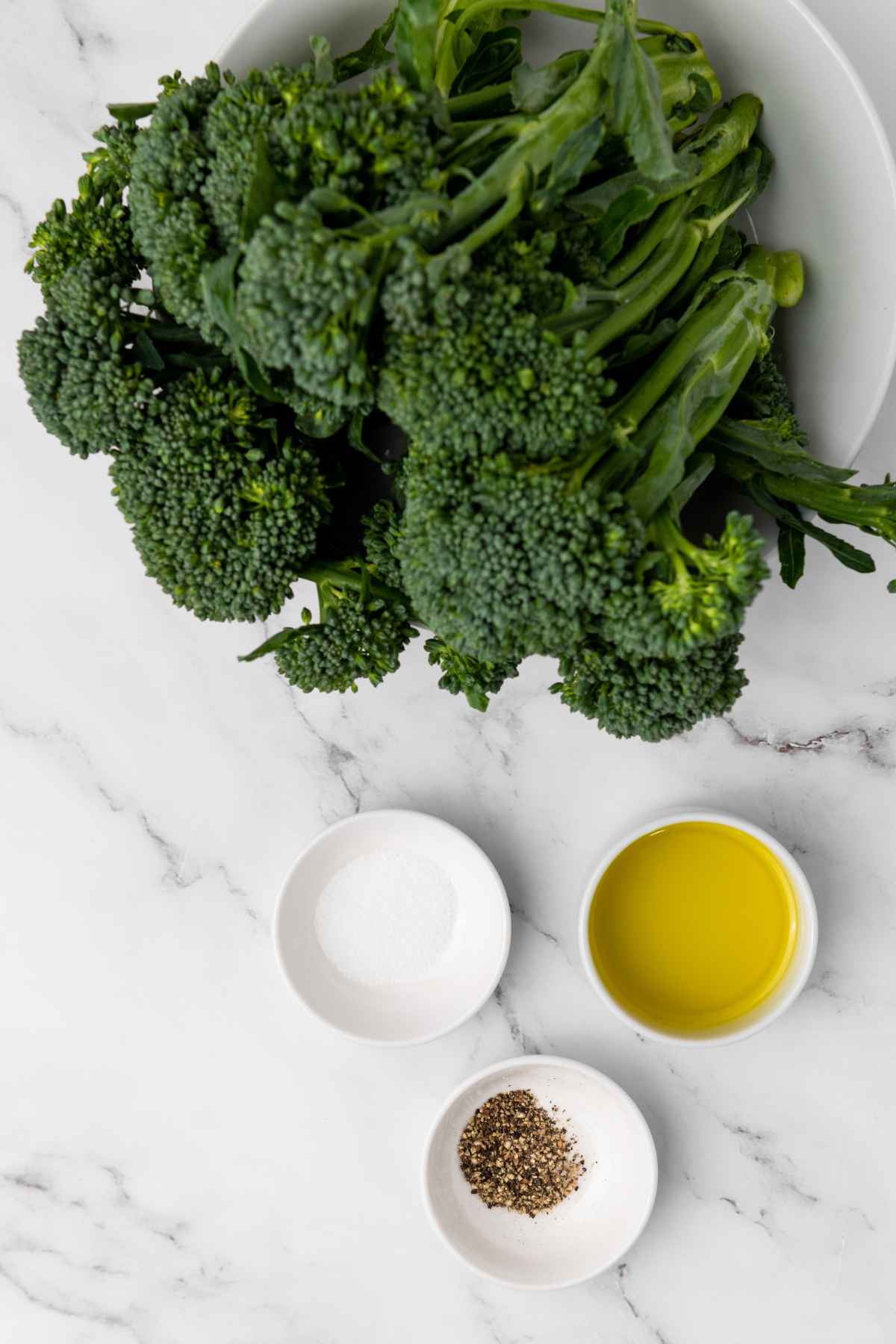 Raw broccolini, salt, pepper, and olive oil on a marbled surface.