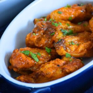 Buffalo chicken wings coated in a bright orange sauce, served in a white ceramic baking dish with a blue handle, and garnished with chopped fresh herbs.