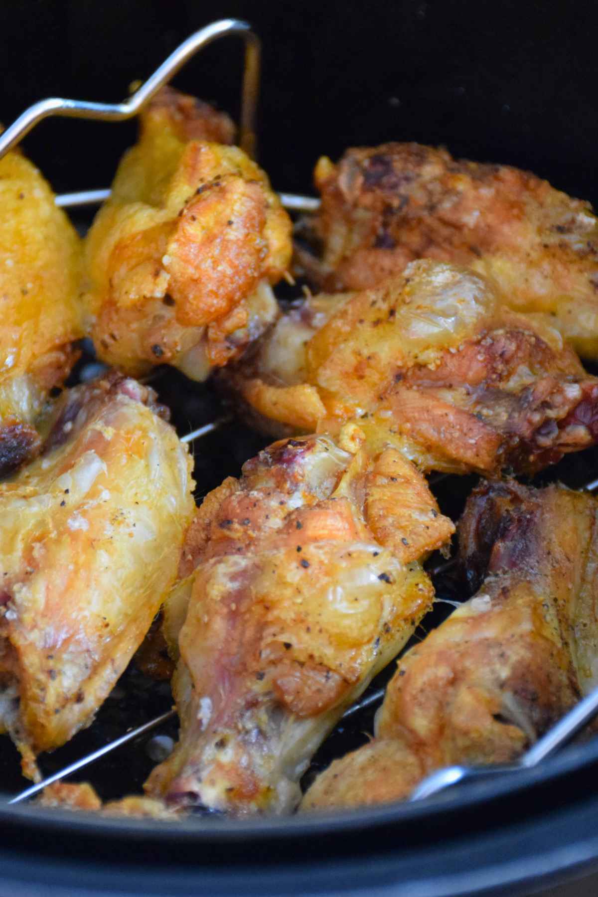 Close up of crispy, golden brown chicken wings cooking in an air fryer basket, seasoned lightly with salt and pepper, with visible grill rack inside the fryer.