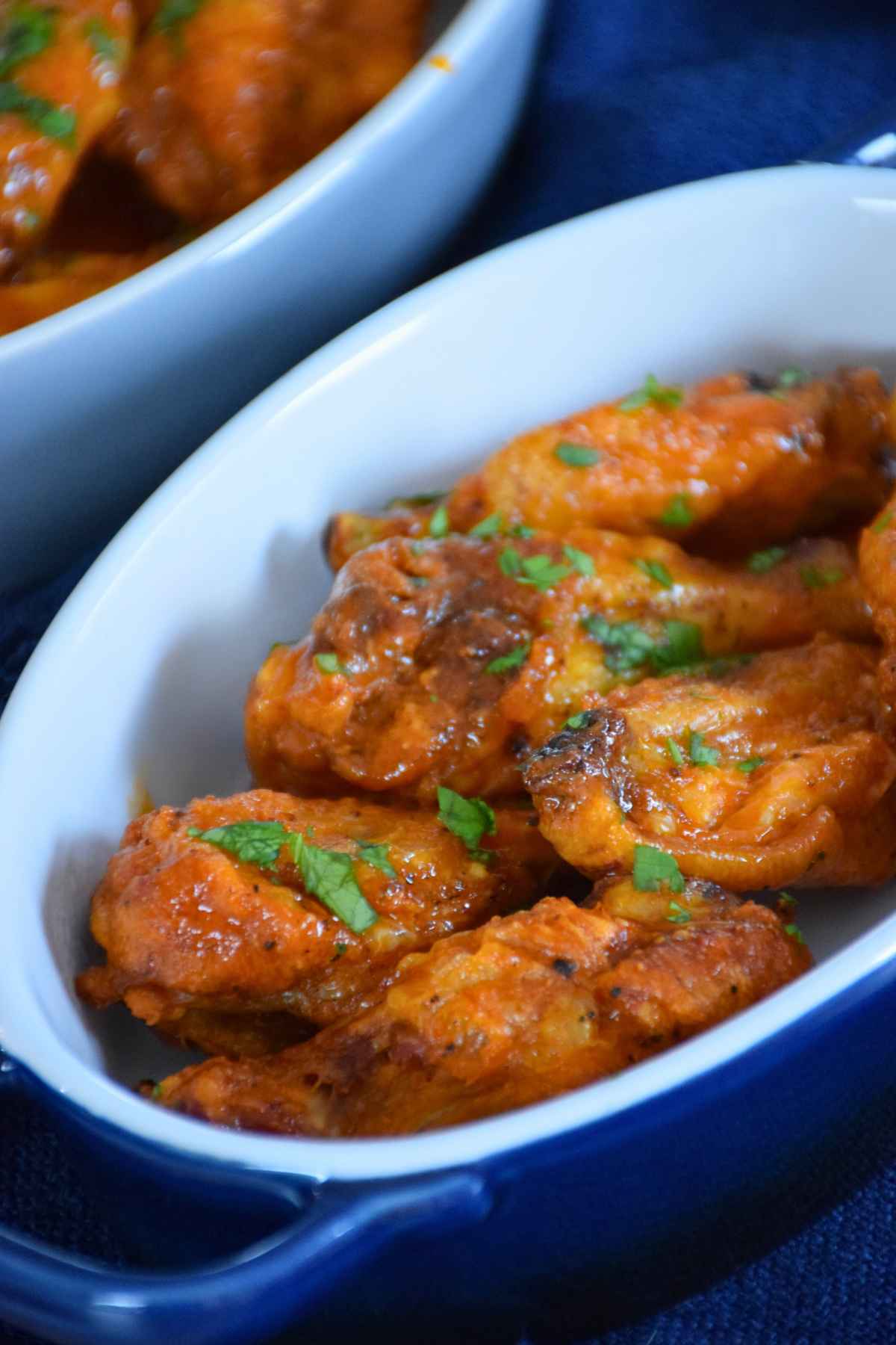 Buffalo chicken wings coated in a bright orange sauce, served in a white ceramic baking dish with a blue handle, and garnished with chopped fresh herbs.