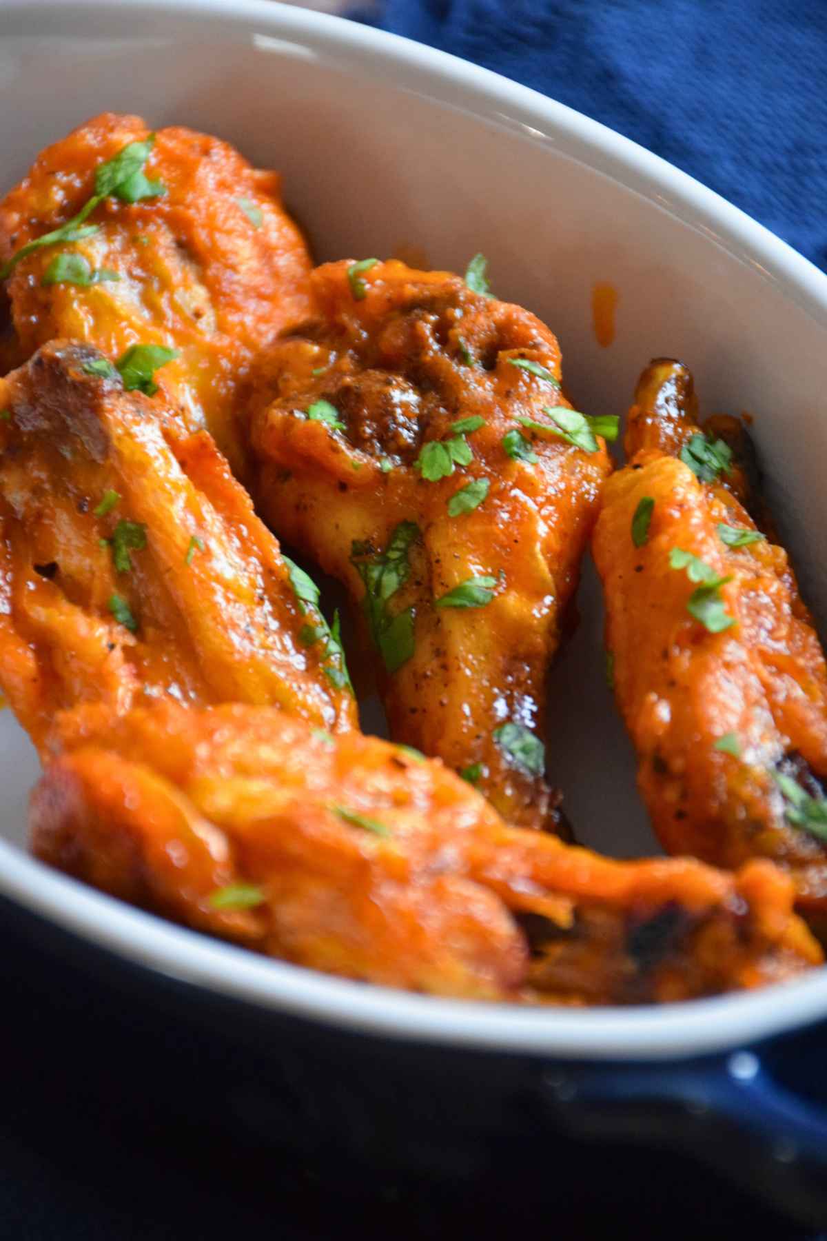Buffalo chicken wings coated in a bright orange sauce, served in a white ceramic baking dish with a blue handle, and garnished with chopped fresh herbs.