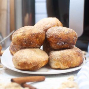 plate stacked with golden brown pumpkin pie bombs coated in cinnamon sugar, with an air fryer in the background