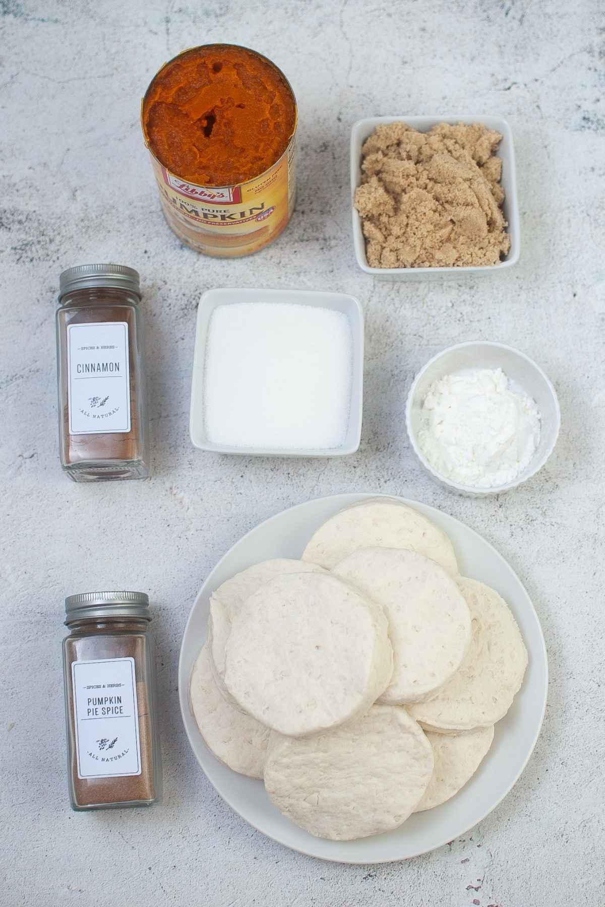 Ingredients laid out on white marble surface including canned pumpkin, brown sugar, white sugar, spices, and biscuit dough.