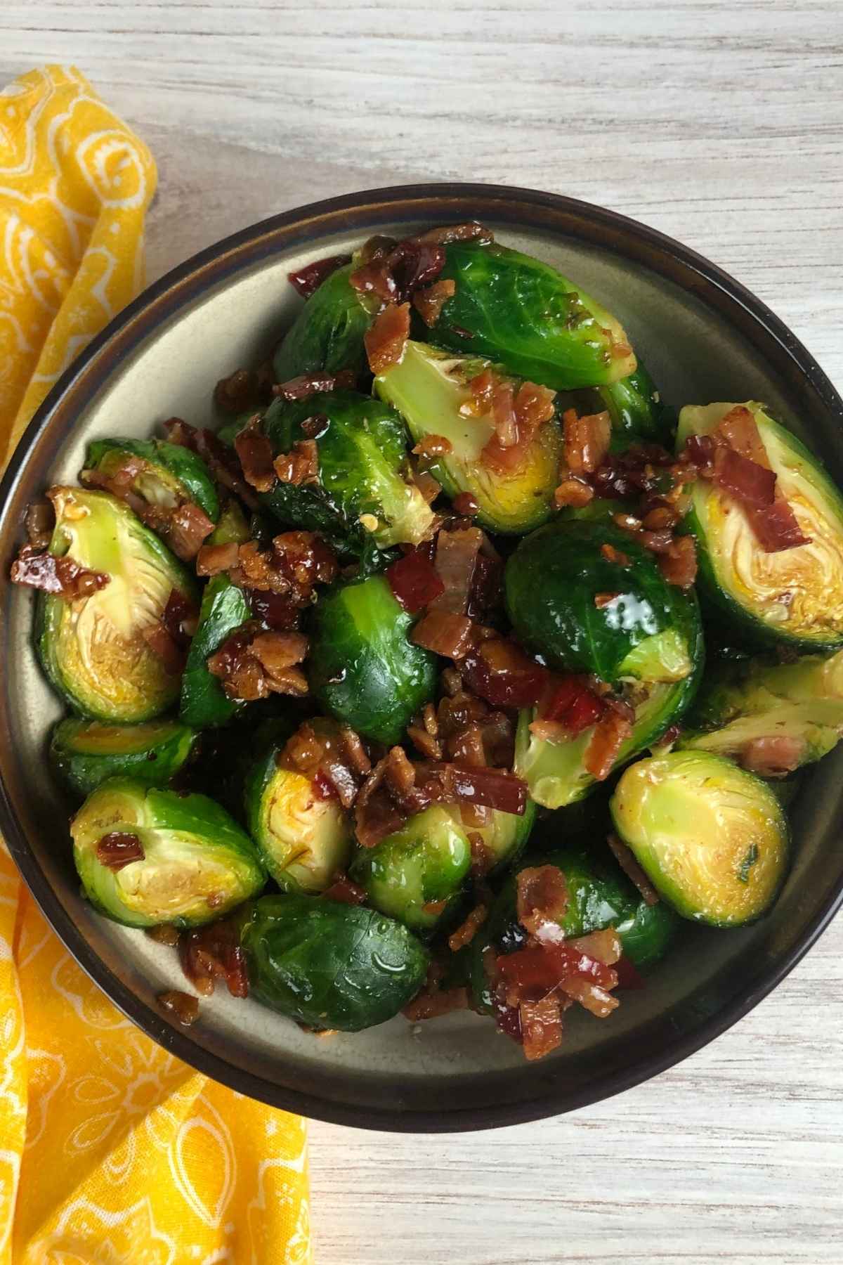 A brown and cream serving bowl filled with crispy air-fried Brussels sprouts mixed with crumbled bacon, coated in glossy pepper jelly.