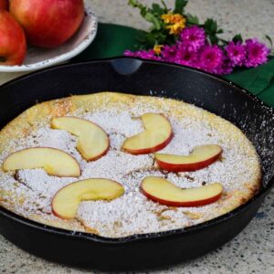Apple Dutch Baby pancake in a cast iron skillet, dusted with powdered sugarn and topped with fresh apple slices, with flowers in the background.