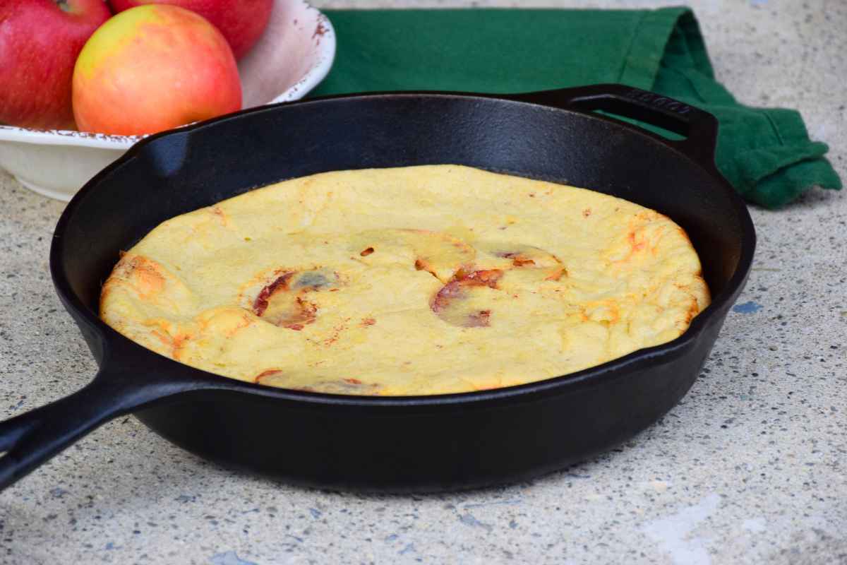Baked apple dutch baby pancake in a cast iron skillet, plain with no toppings, with a bowl of apples in the background.