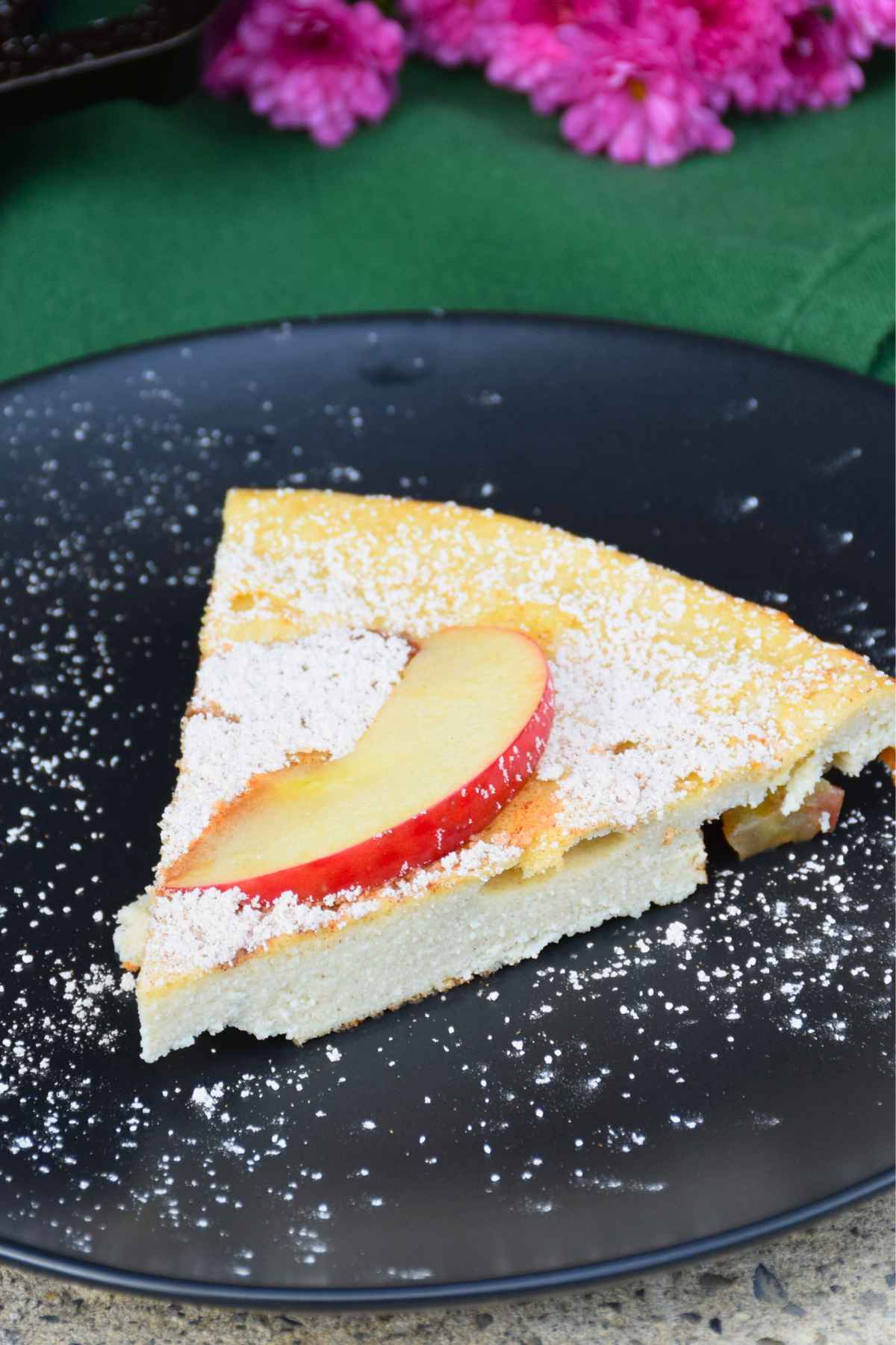Slice of Apple Dutch Baby Pancake on a black plate, dusted with powdered sugar and topped with a fresh apple slice.