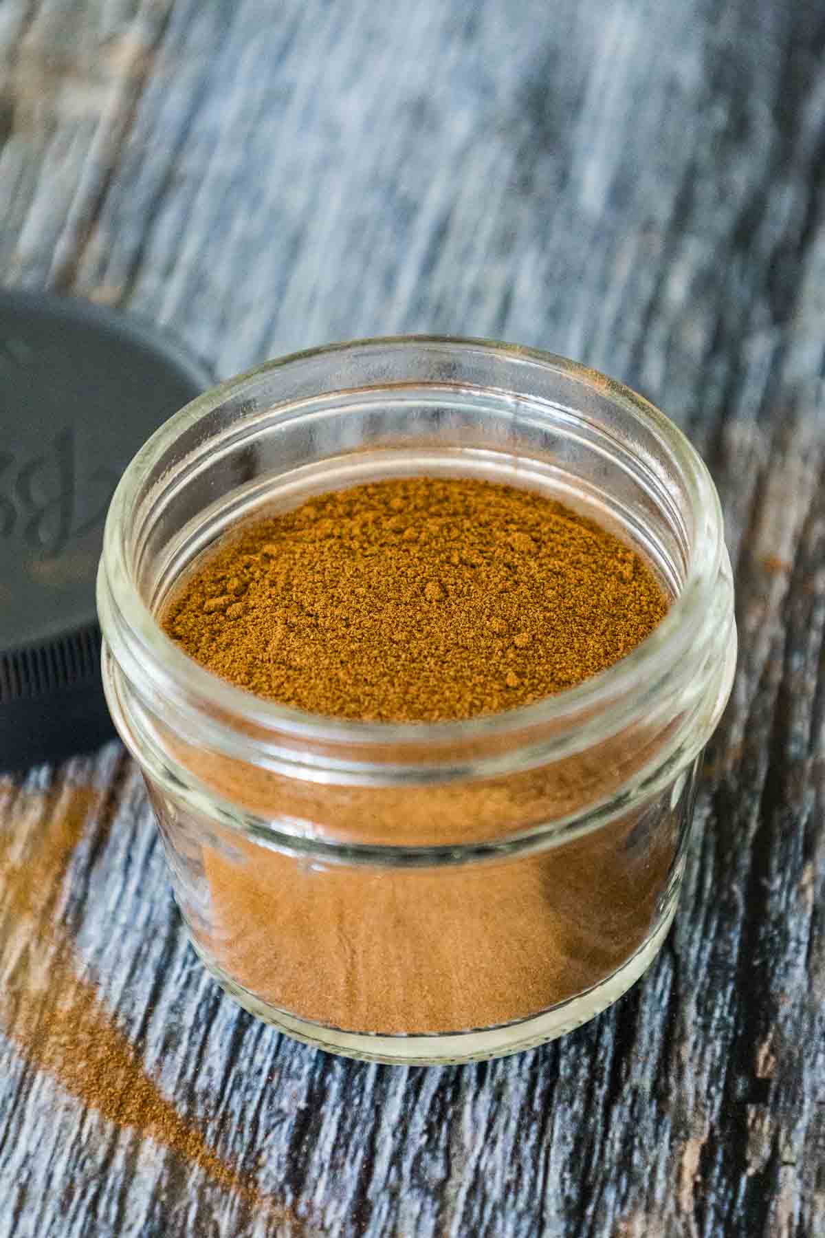 A small glass jar filled with apple pie spice, sitting on a wooden surface with the lid beside it.