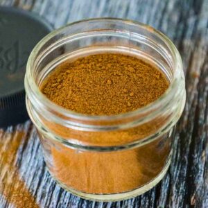 A small glass jar filled with apple pie spice, sitting on a wooden surface with the lid beside it.