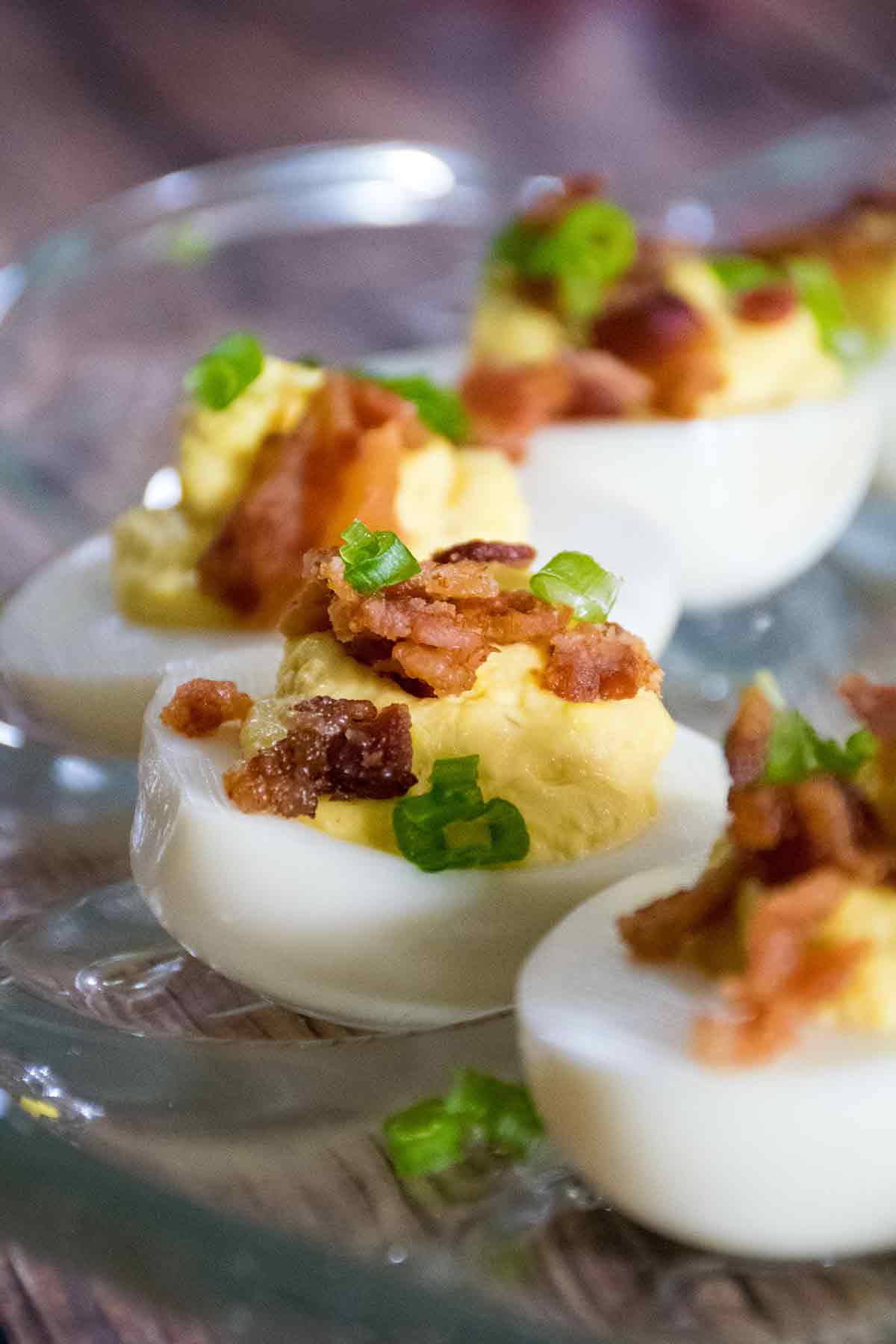 Row of deviled eggs on a glass tray, filled with creamy yolk mixture and garnished with bacon bits and green onion slices.