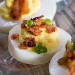 Close up of a bacon deviled egg topped with crumbled bacon and chopped chives on a wooden surface.
