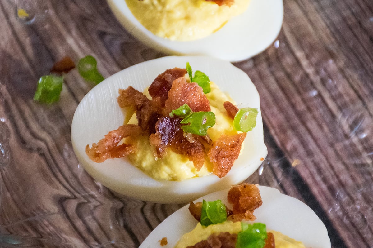 Close up of a deviled egg topped with crumbled bacon and chopped chives on a wooden surface.