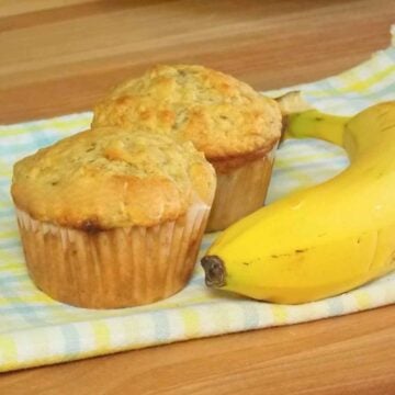 Two whole golden baked banana muffins with a whole banana to the side on a patterned towel.