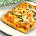 A single square slice of buffalo chicken flatbread on a white plate, garnished with melted cheese and chopped parsley with a green patterned napkin in the background.