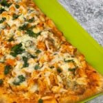 Close up of a fully baked flatbread pizza topped with melted cheese, chunks of chicken, and fresh herbs on a bright green cutting board.