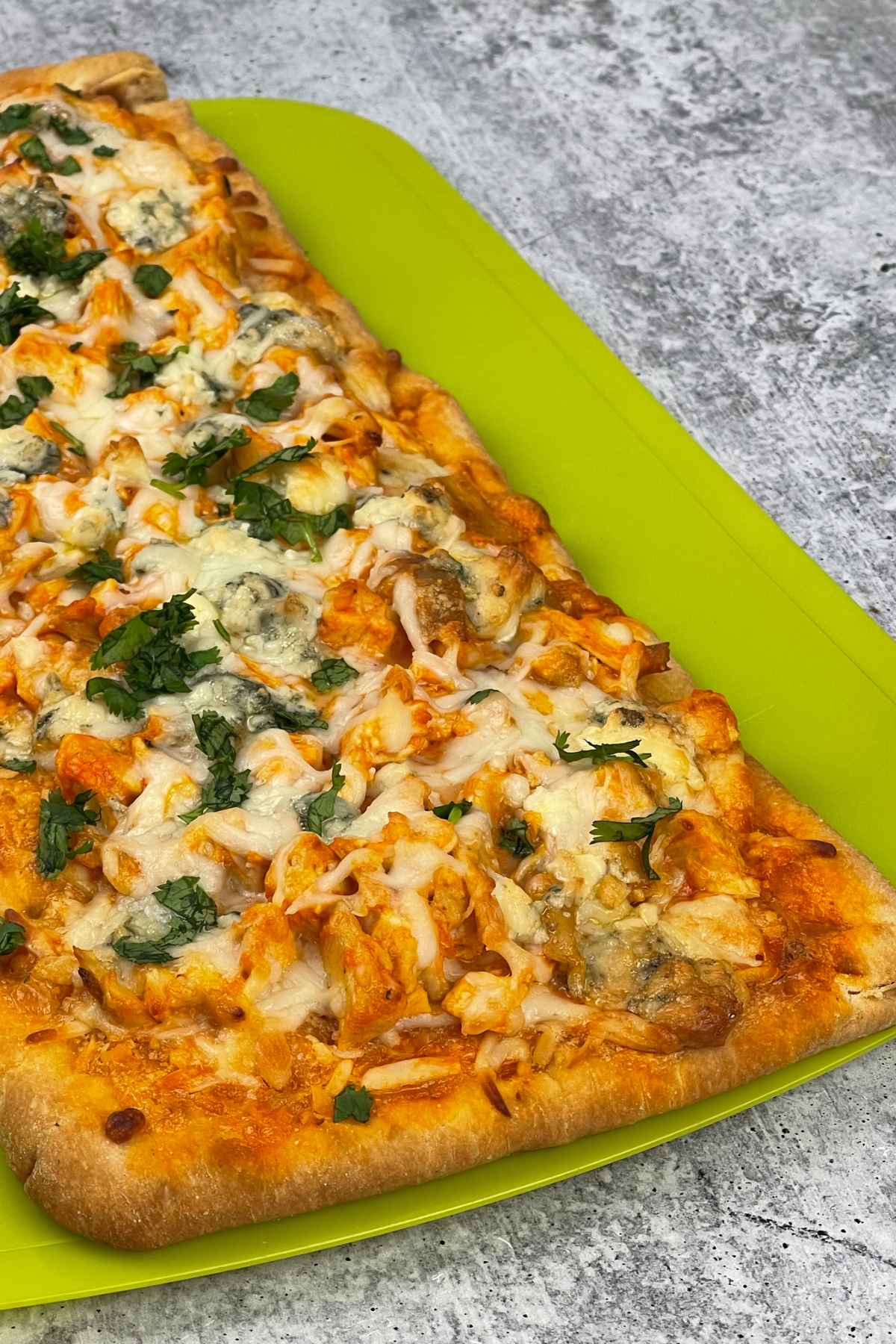 Close up of a fully baked flatbread pizza topped with melted cheese, chunks of chicken, and fresh herbs on a bright green cutting board.