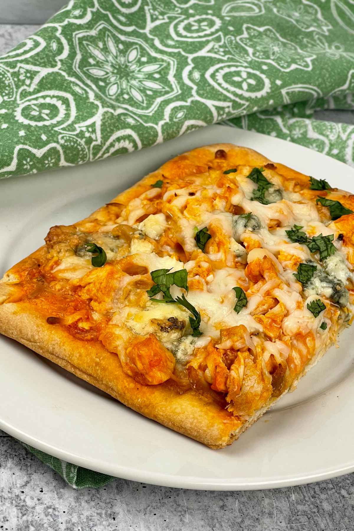 A single square slice of buffalo chicken flatbread on a white plate, garnished with melted cheese and chopped parsley with a green patterned napkin in the background.