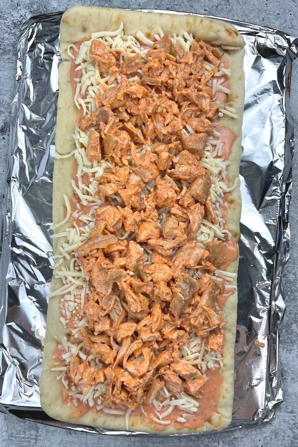 Unbaked flatbread topped with shredded mozzarella cheese, generous layer of buffalo chicken, set on foil over a baking sheet.