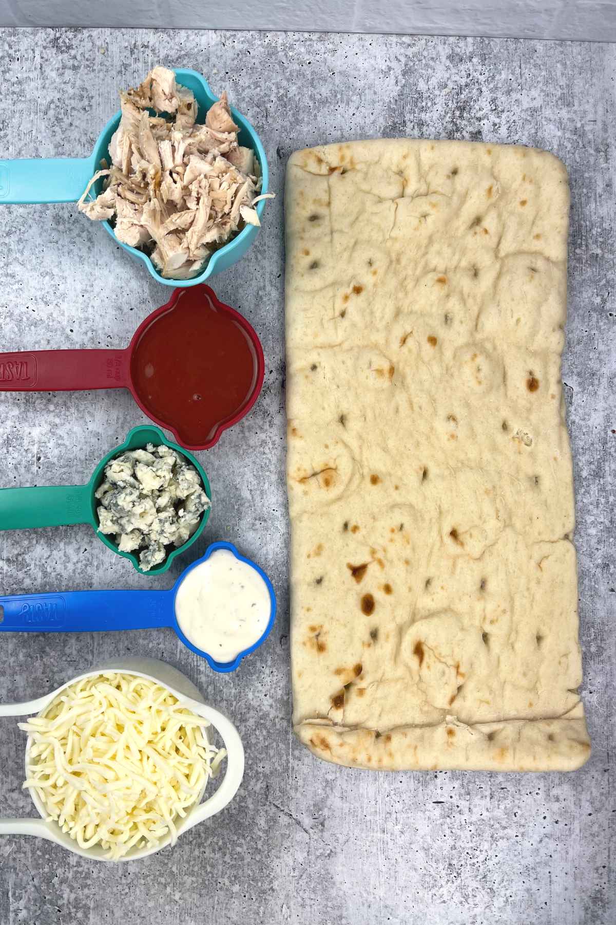 Overhead view of measured ingredients including shredded chicken, buffalo sauce, blue cheese, ranch dressing, shredded mozzarella, and an oblong flatbread.
