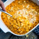 A large white casserole dish with buffalo chicken mac and cheese with a hand spooning out a serving.