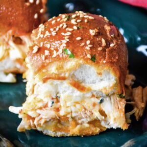 Close up of a buffalo chicken slider with a glossy, golden-brown bun topped with sesame seeds and chopped parsley, revealing shredded chicken and melted cheese inside.
